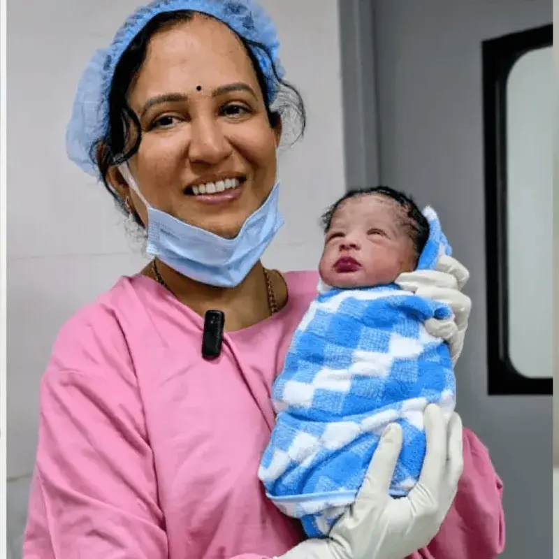 Dr. Padmaja in a pink gown and surgical cap, smiling as she holds a newborn wrapped in a blue and white checked blanket