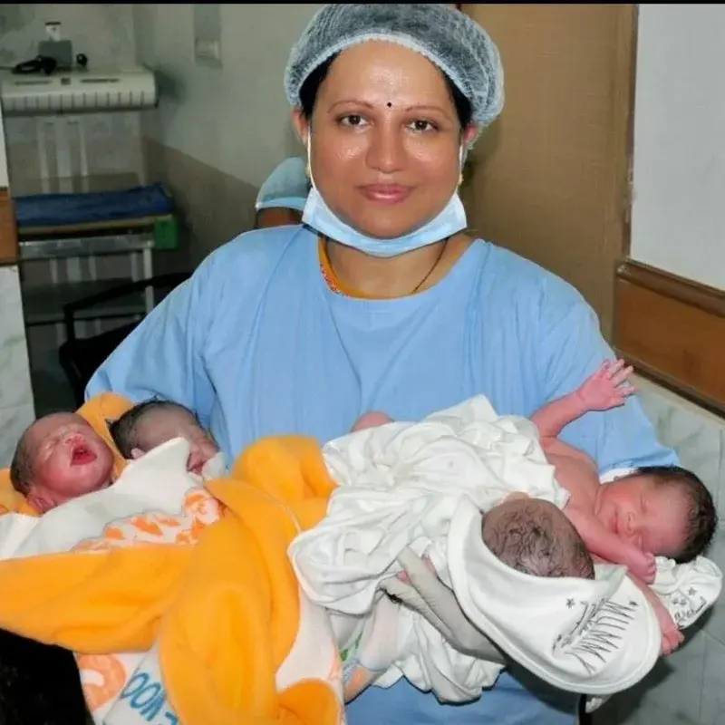 Dr. Padmaja in blue scrubs holding four newborn babies wrapped in yellow and white blankets