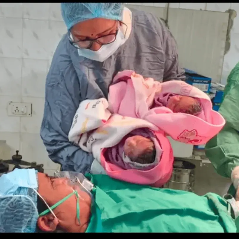 A member of the clinical team showing newborn twins in pink blankets to their mother in theatre
