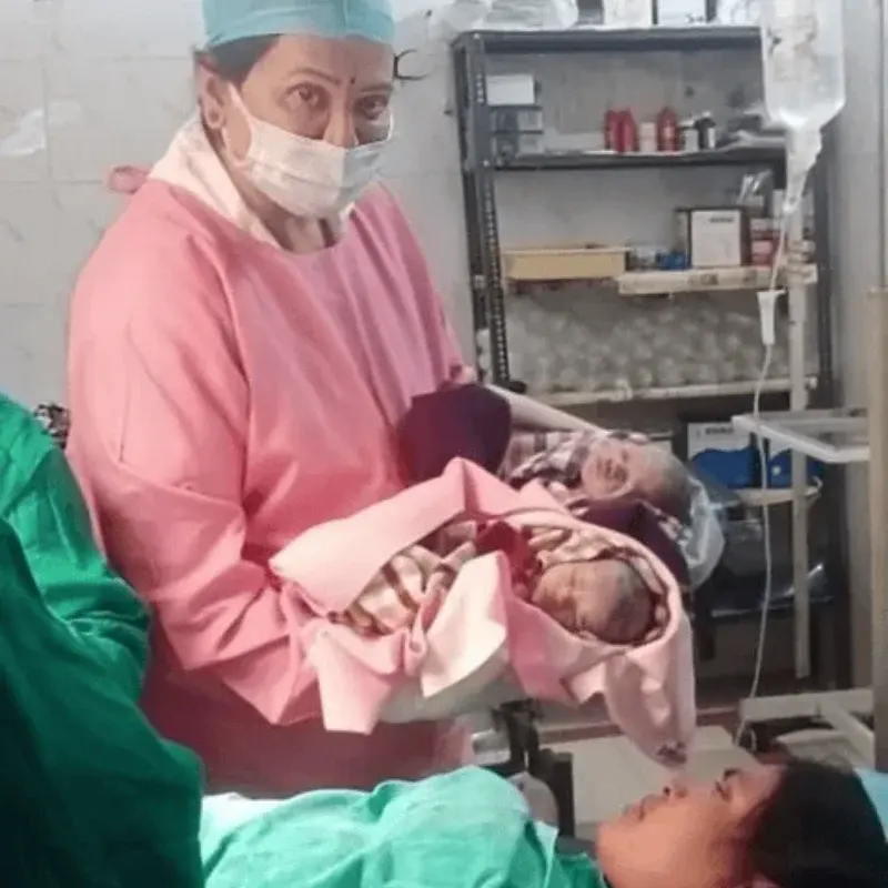A member of the care team in a pink gown holding newborn twins beside their mother after delivery